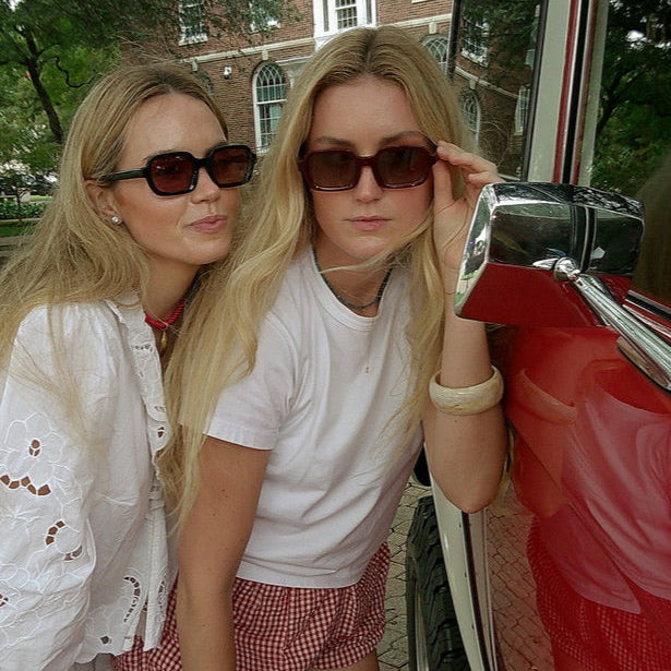 Two women posing next to a red vehicle with sunglasses on, outdoors.