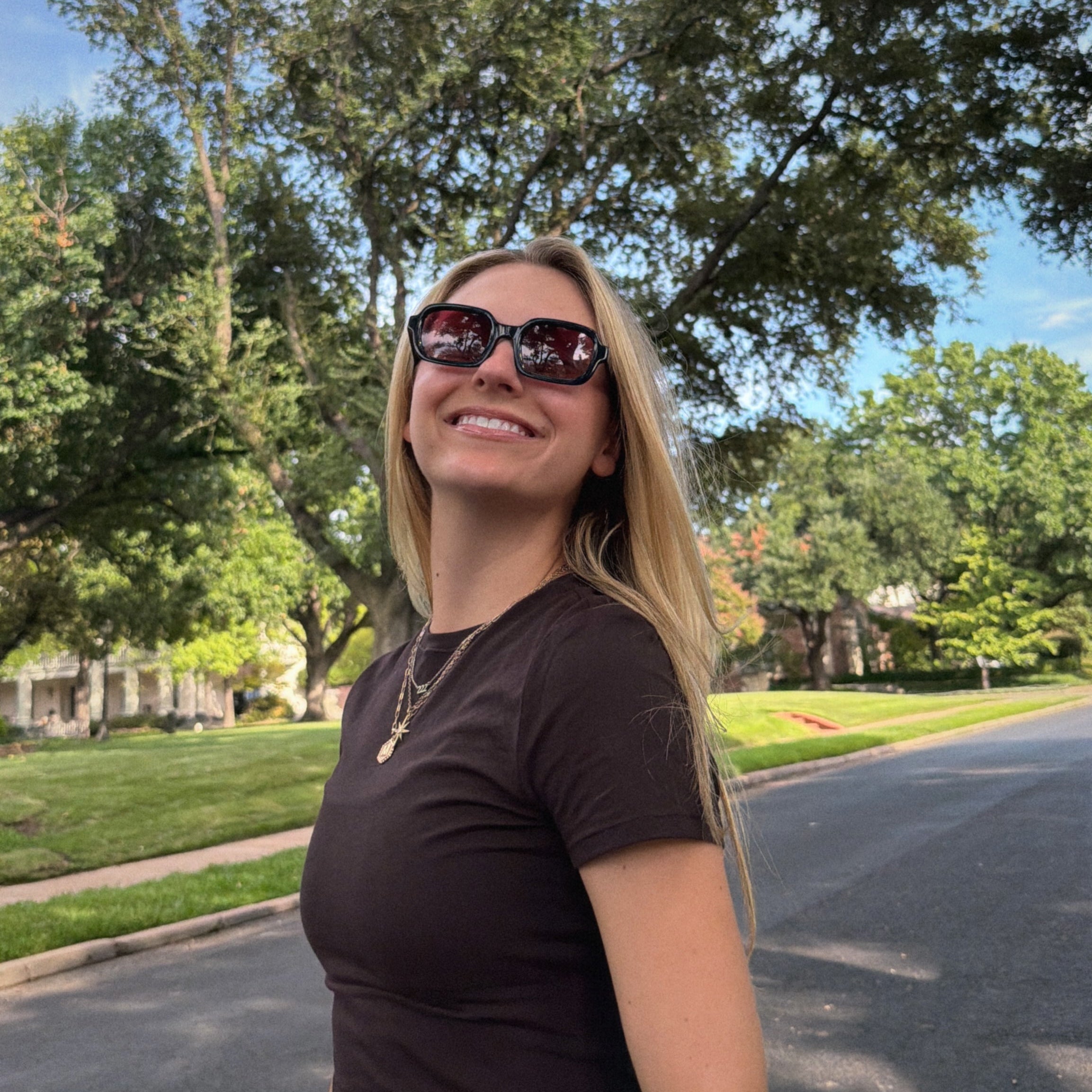 Woman wearing sunglasses and a brown shirt standing on a street with trees and grass in the background