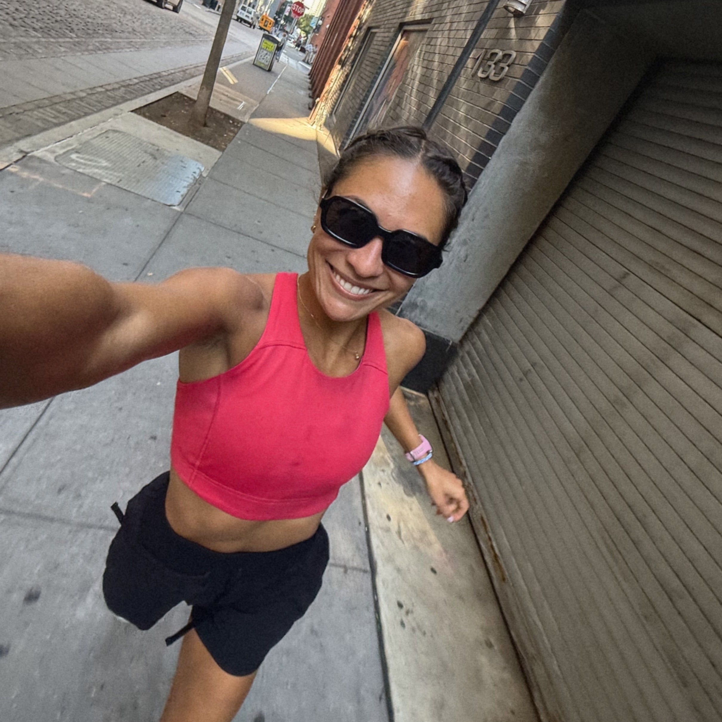 Woman in pink sports bra and black shorts taking a selfie on a city sidewalk.