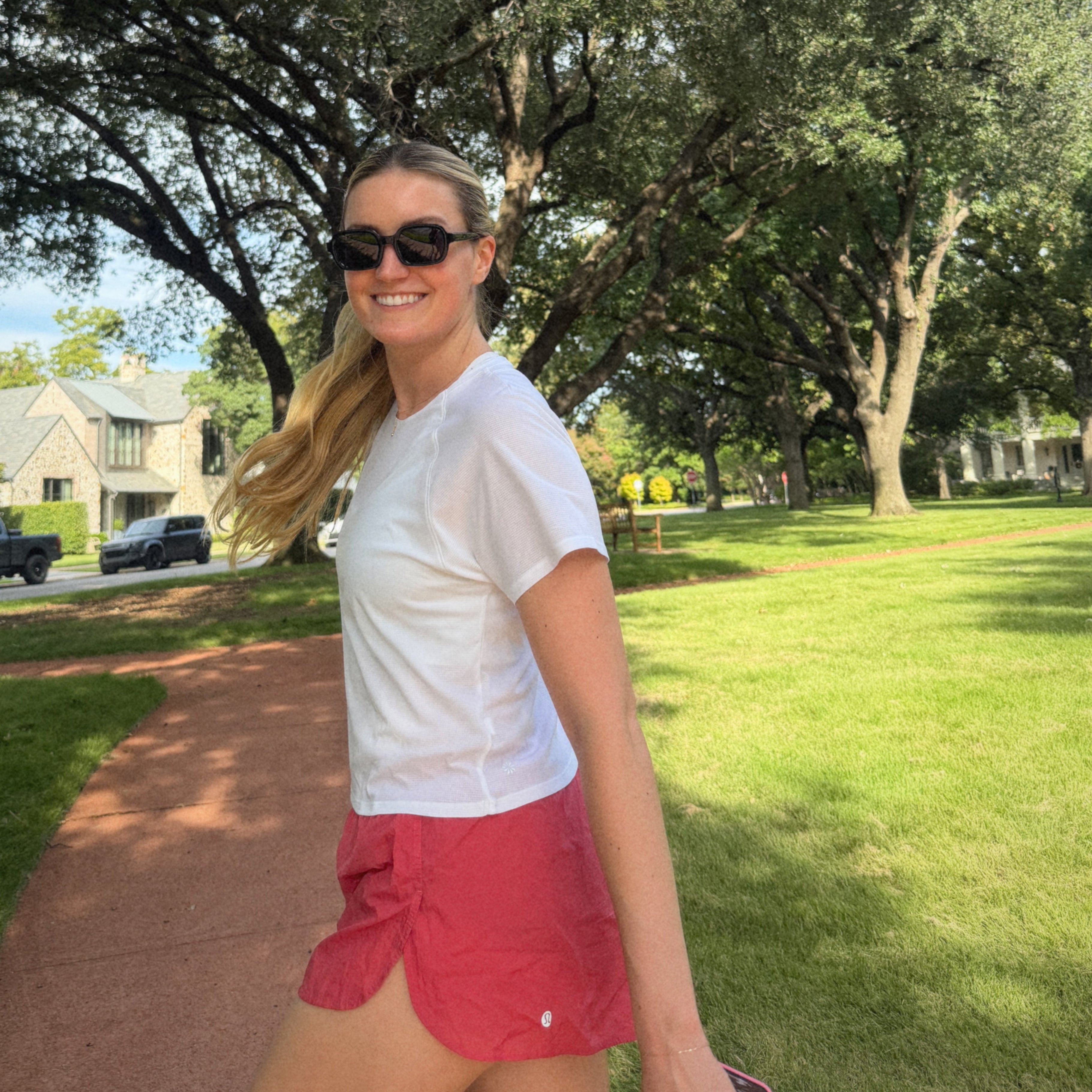 Woman in white shirt and red shorts standing on a sidewalk with trees and houses in the background