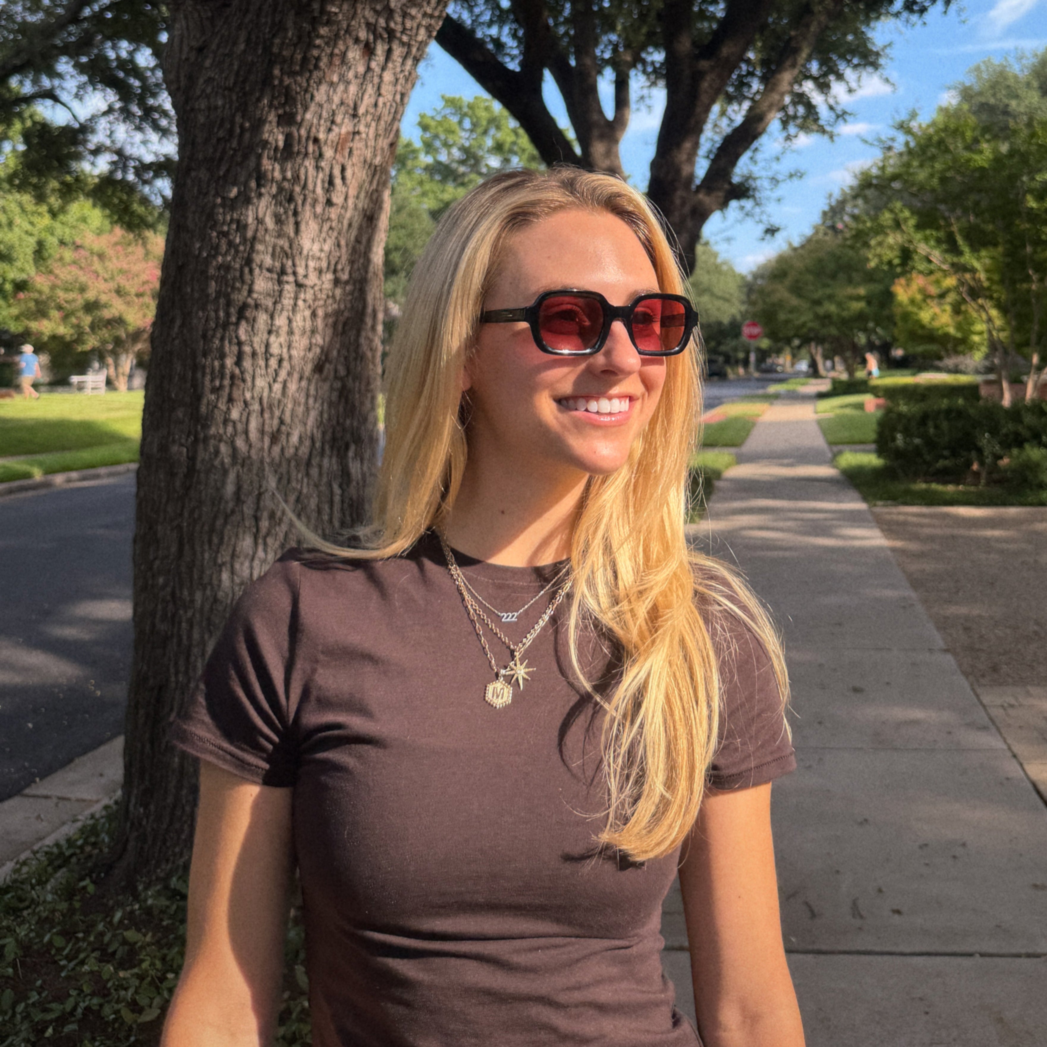 Woman wearing sunglasses and a brown shirt standing on a sidewalk with trees and grass in the background