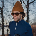 Man wearing a blue jacket, brown beanie, and sunglasses outdoors with trees in the background