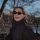Person wearing sunglasses and a dark coat standing outdoors with trees and water in the background