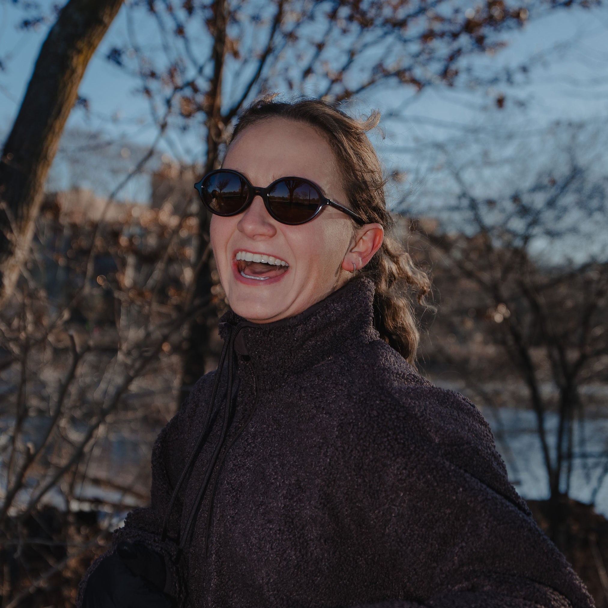 Person wearing sunglasses and a dark coat standing outdoors with trees and water in the background