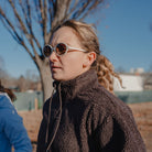 Person wearing a brown jacket and sunglasses outdoors with a blurred background