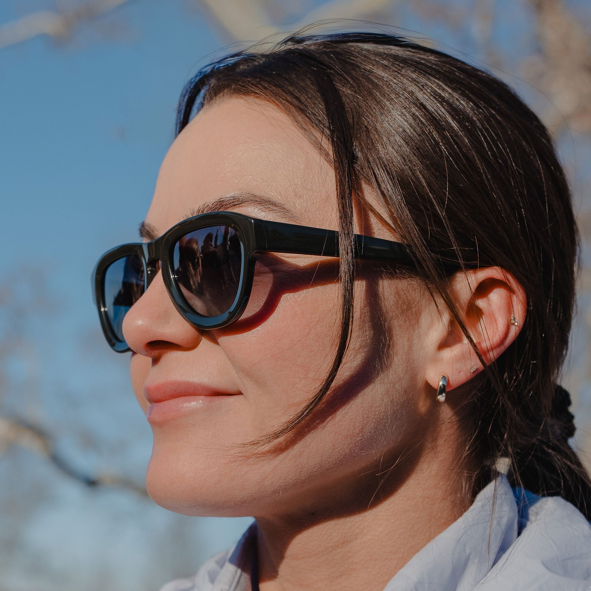 Woman wearing sunglasses and a light gray jacket outdoors with a clear blue sky.
