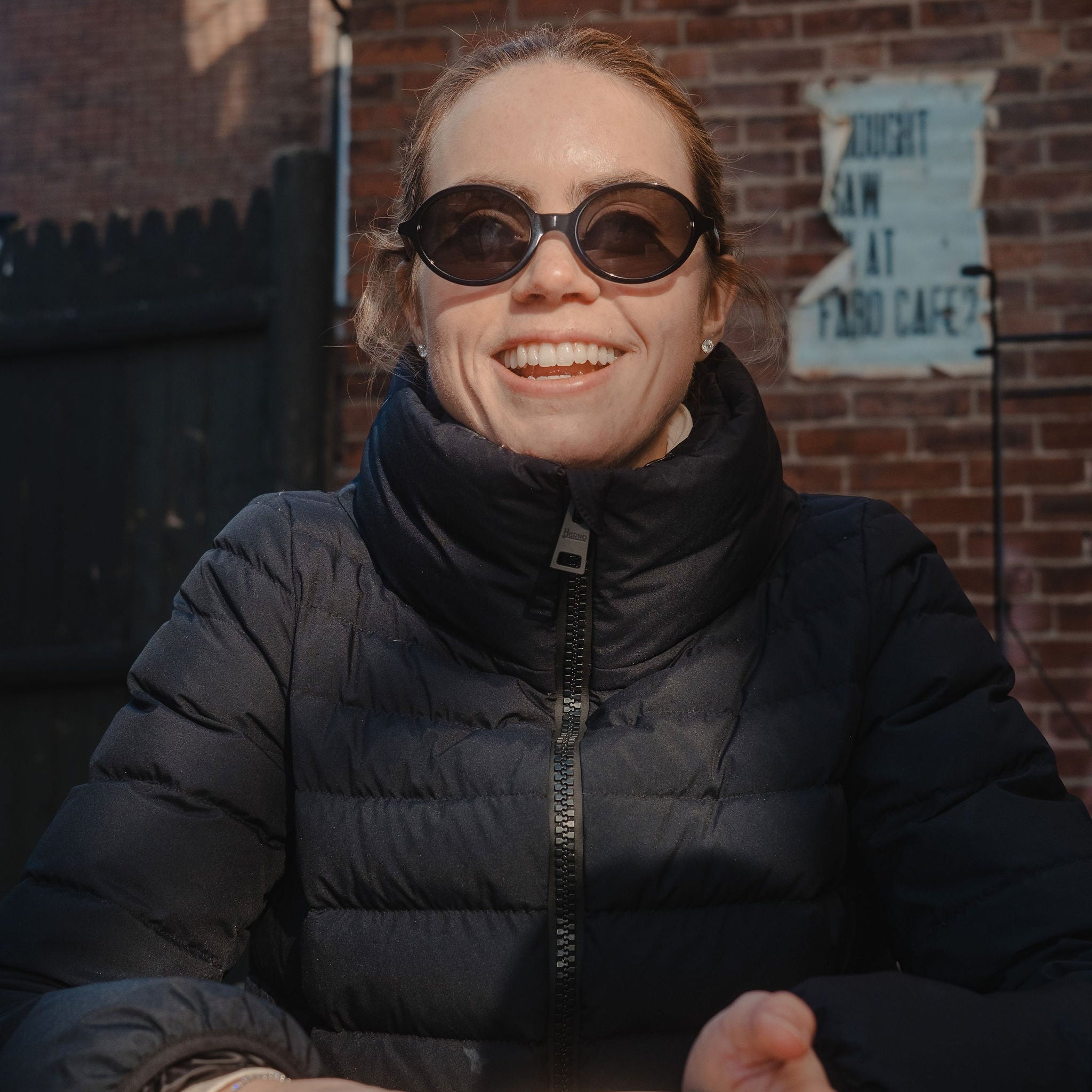 Person wearing sunglasses and a black puffer jacket holding a cup outdoors.