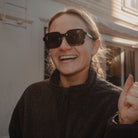 Person wearing sunglasses and a dark jacket, giving a thumbs-up in front of a building.