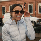 Woman wearing sunglasses and a light blue puffer jacket in an urban setting.