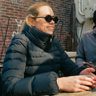 Person wearing sunglasses and a dark jacket sitting outdoors near a brick wall.