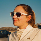 Woman wearing sunglasses and a light-colored jacket with a logo, standing outdoors.