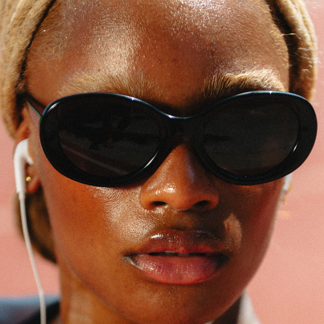 A close-up lifestyle shot of a woman wearing black cat-eye sunglasses, white wired earphones, and gold necklaces, with a warm pinkish-orange background.