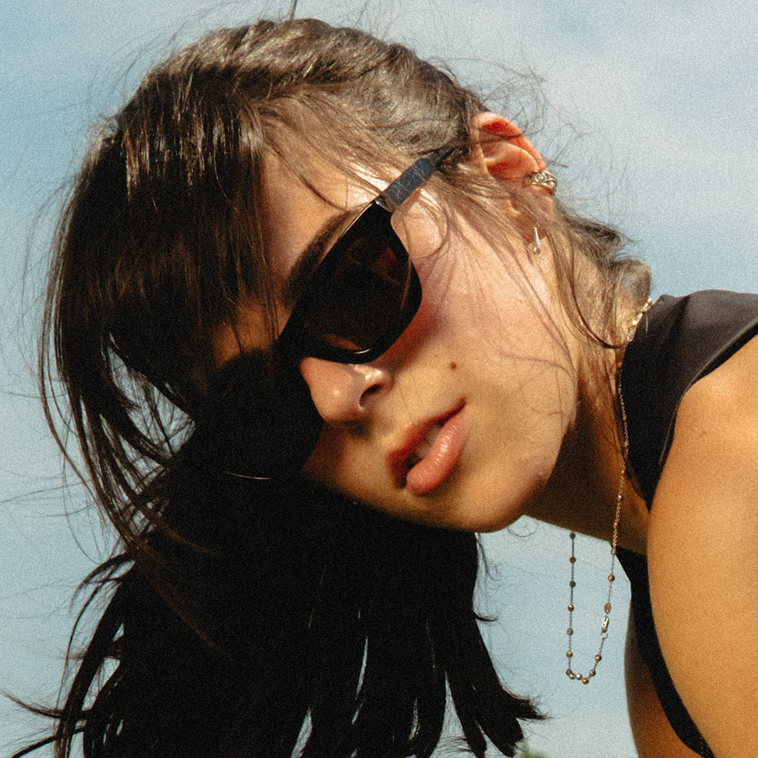A close-up lifestyle shot of a woman with dark hair wearing black sunglasses, a delicate beaded necklace, and a black top, set against a bright sky with green trees in the background.