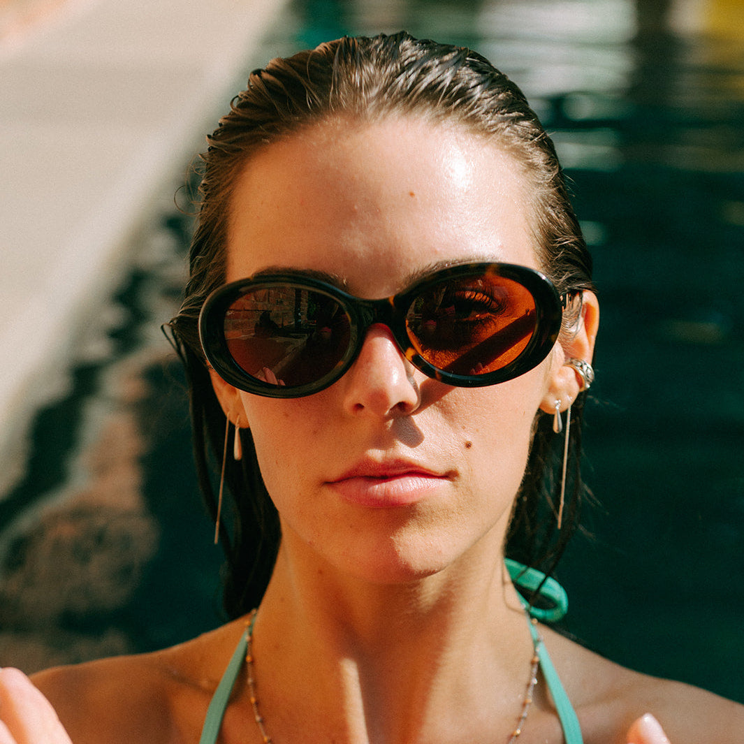 A woman wearing dark sunglasses and a turquoise bikini top holds a large slice of watermelon in front of her face, with a poolside background.