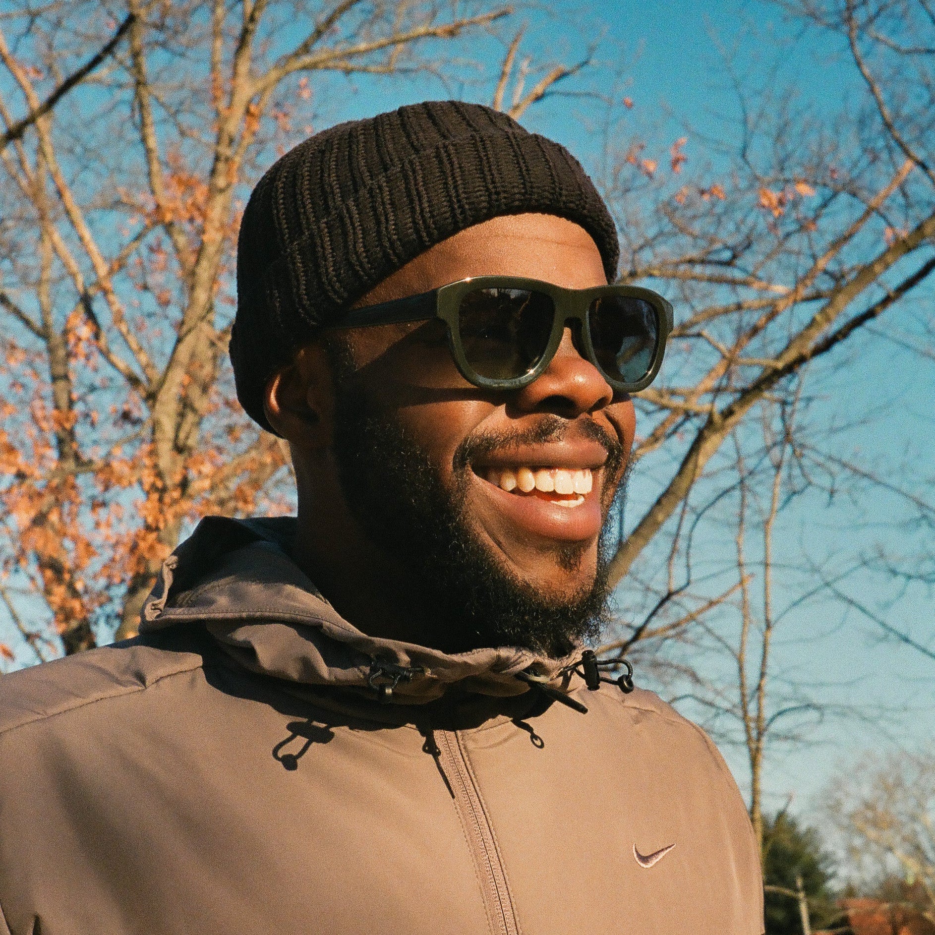 Man wearing sunglasses and a beanie with a clear blue sky and trees in the background